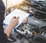 Une personne écrit sur un presse-papiers tout en inspectant le moteur d'une voiture, s'assurant que les révisions et les vidanges sont effectuées avec précision.