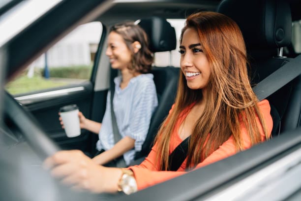Deux femmes sont assises dans une voiture, toutes deux attachées à leur ceinture de sécurité. La femme assise à la place du conducteur est souriante et concentrée sur la conduite, réfléchissant peut-être à ses récentes révisions de vidange moteur, tandis que le passager tient une tasse de café et rit.